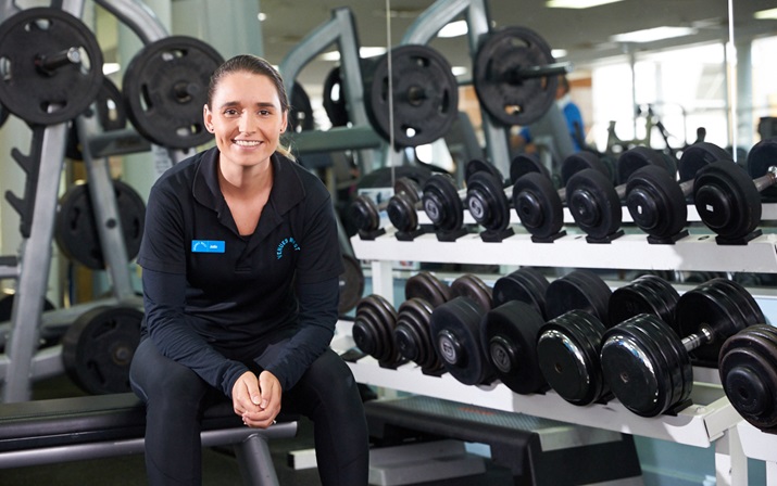 Gym Staff A photo of a gym staff member sitting on a bench next to a rack of free weights