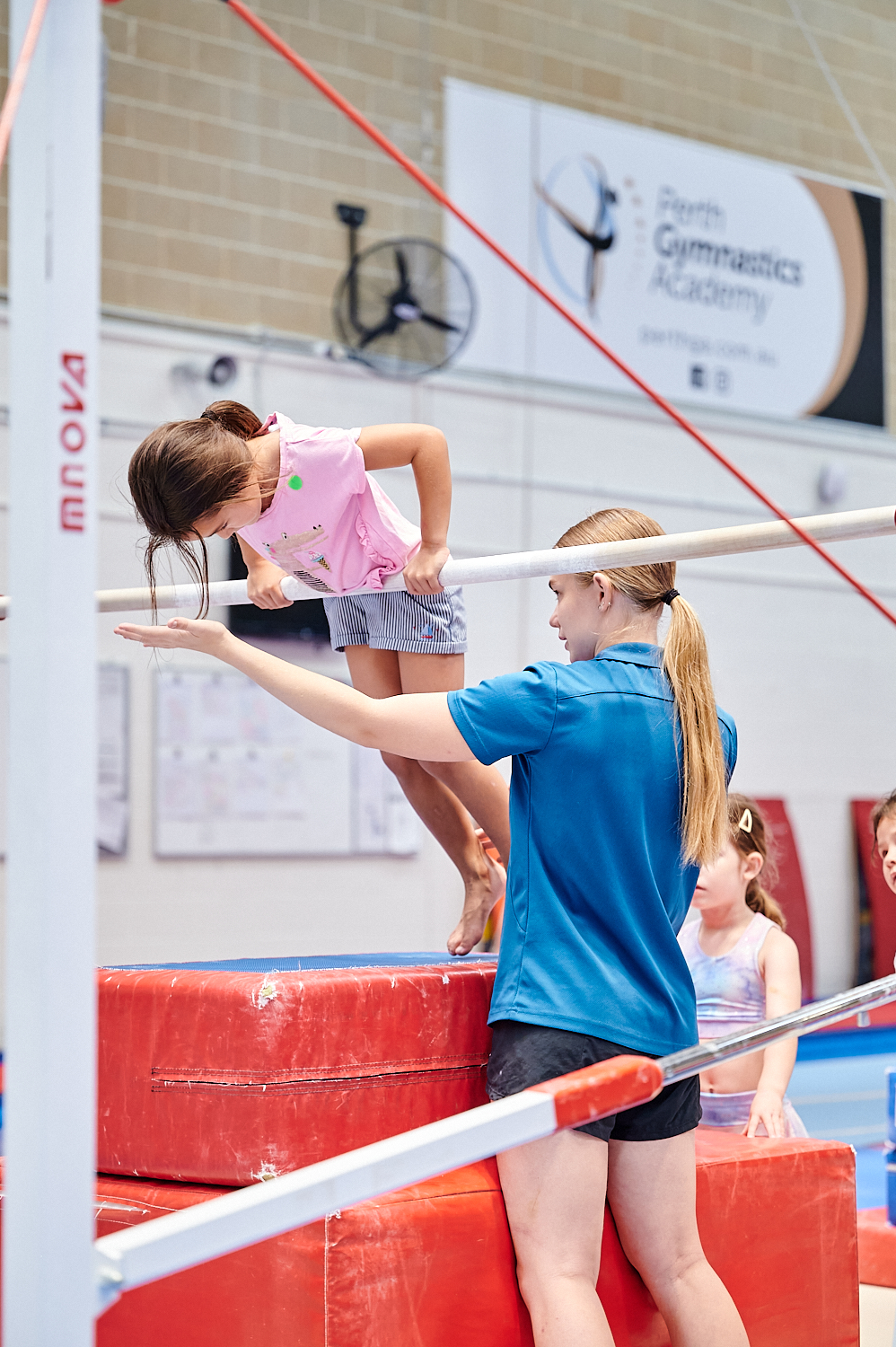 A child pulling herself up using a gymnastics bar with the assistance of a teacher at the Perth HPC Kids Gymnastics in Mount Claremont, Perth