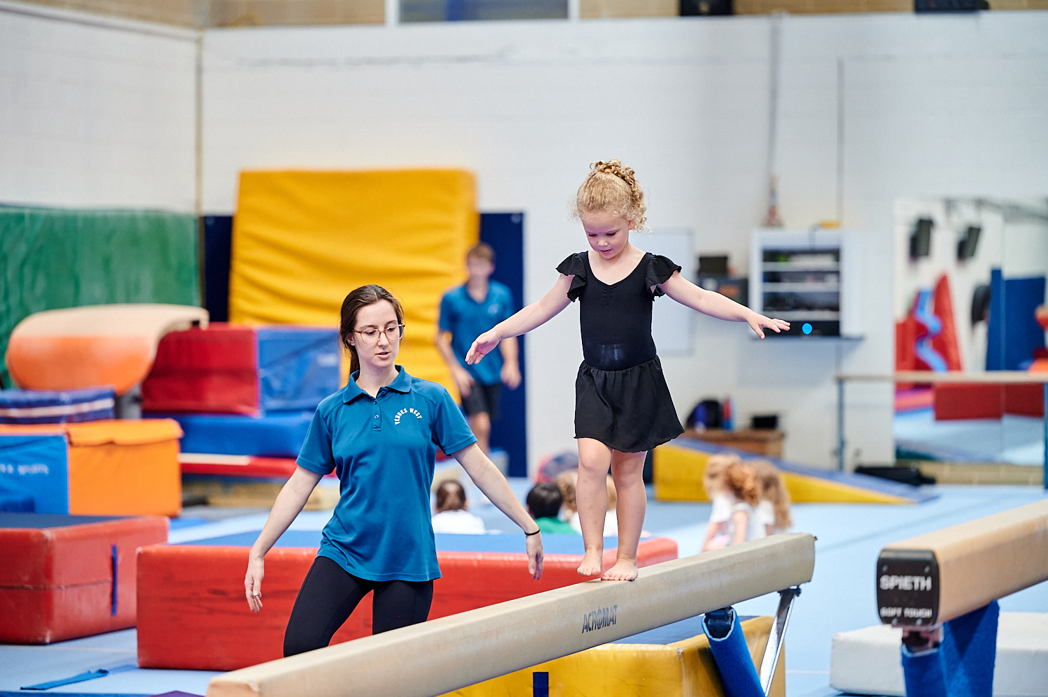 A child walking along a balance beam with a teacher assisting at the Perth HPC Kids Gymnastics in Mount Claremont, Perth