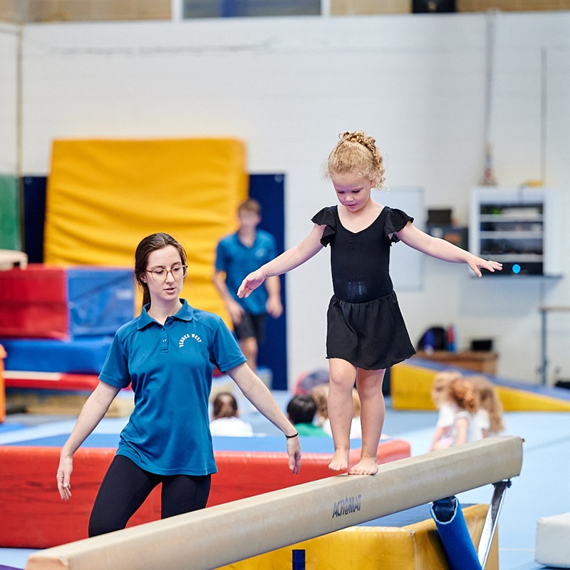 Young girl balancing on beam with assistance from Gymnastics Instructor at Kids Gym class at Perth HPC, Mount Claremont, Perth